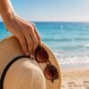 Mano con manicure rosa naturale che tiene un cappello di paglia e occhiali da sole sulla spiaggia, immagine che rappresenta i fattori estivi che possono influenzare la durata della ricostruzione delle unghie