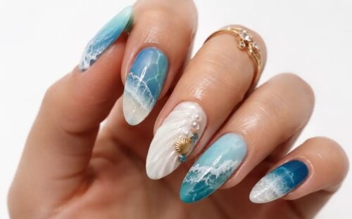 Mano con nail art effetto mare realistica, sfumature azzurre e blu, conchiglia tridimensionale e dettagli che ricordano la schiuma delle onde su sfondo bianco.